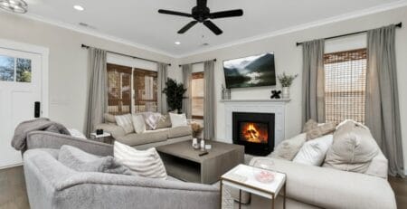 Cozy modern living room with neutral-toned sectional sofas around a low coffee table, a lit fireplace with a TV mounted above, a ceiling fan overhead, and windows with bamboo shades and gray curtains.