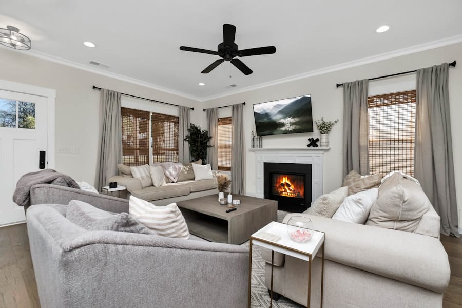 Cozy modern living room with neutral-toned sectional sofas around a low coffee table, a lit fireplace with a TV mounted above, a ceiling fan overhead, and windows with bamboo shades and gray curtains.