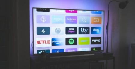 Smart TV in a dim living room displaying a grid of streaming app icons (e.g., Netflix, Prime Video, Spotify, BBC iPlayer) with a purple backlight and a floor lamp beside it.