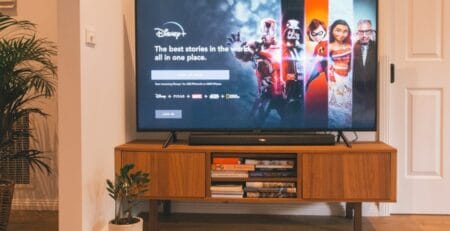 Living room scene with a flat-screen TV on a mid-century wooden console displaying the Disney+ sign-in screen with character art, books stored on the shelf below, and a potted plant beside.