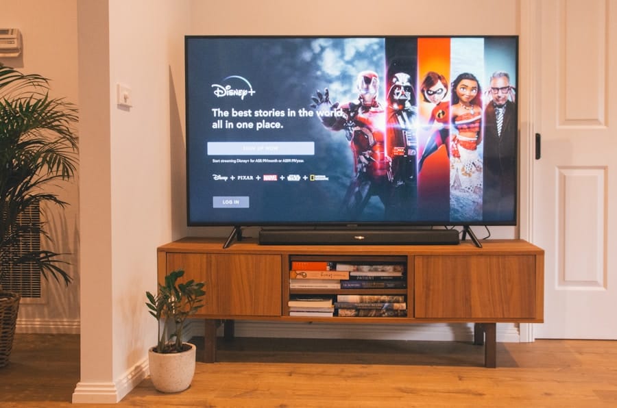 Living room scene with a flat-screen TV on a mid-century wooden console displaying the Disney+ sign-in screen with character art, books stored on the shelf below, and a potted plant beside.