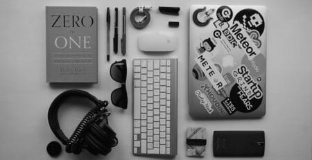 Monochrome flat lay of neatly arranged tech essentials: a sticker-covered laptop, wireless keyboard and mouse, over-ear headphones, pens, sunglasses, cables, SD cards, a smartphone, and the book “Zero to One” on a light desk.