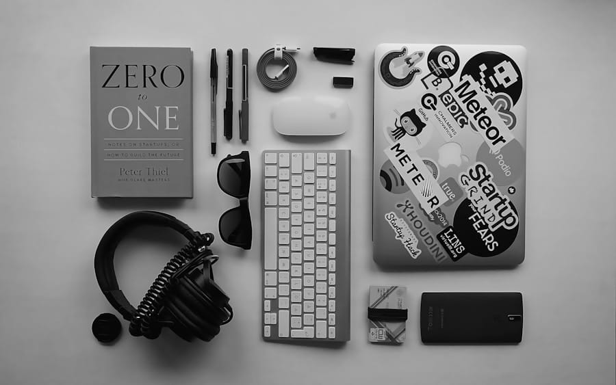 Monochrome flat lay of neatly arranged tech essentials: a sticker-covered laptop, wireless keyboard and mouse, over-ear headphones, pens, sunglasses, cables, SD cards, a smartphone, and the book “Zero to One” on a light desk.