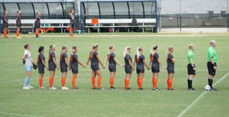 Soccer players in dark gray uniforms with bright orange socks stand hand-in-hand in a line on the field facing two referees before kickoff, with benches and a dugout in the background.