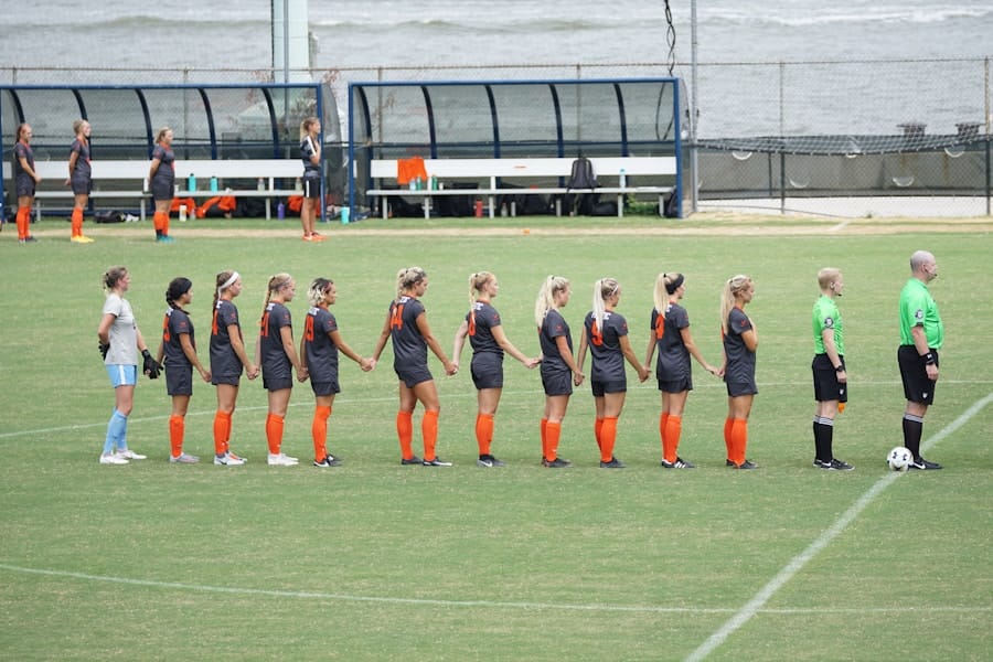 Soccer players in dark gray uniforms with bright orange socks stand hand-in-hand in a line on the field facing two referees before kickoff, with benches and a dugout in the background.