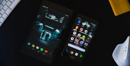 An Android tablet and smartphone on a dark desk, screens on—tablet with a moody city staircase wallpaper and phone showing app icons—beside a white keyboard and ergonomic mouse.