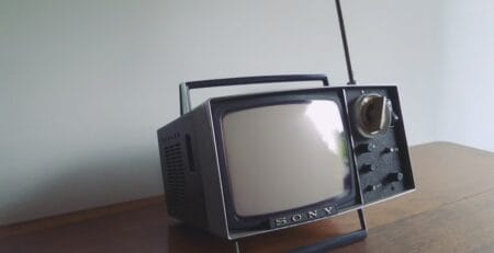 Retro Sony portable CRT television with a telescopic antenna, side knobs, and carry handle, sitting on a wooden table against a plain wall.
