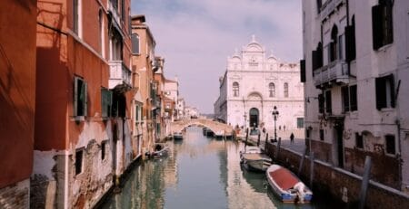 A narrow canal in Venice with moored boats, pastel buildings on both sides, an arched stone footbridge, and a white church facade in the distance under a hazy sky.