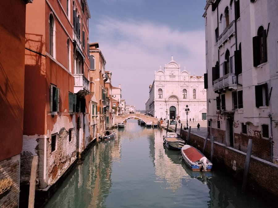A narrow canal in Venice with moored boats, pastel buildings on both sides, an arched stone footbridge, and a white church facade in the distance under a hazy sky.