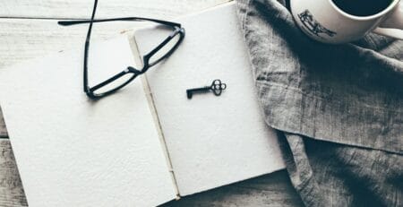 Overhead view of an open blank notebook with black glasses and a small vintage key on its pages, next to a white coffee mug on gray linen atop a rustic wooden table.