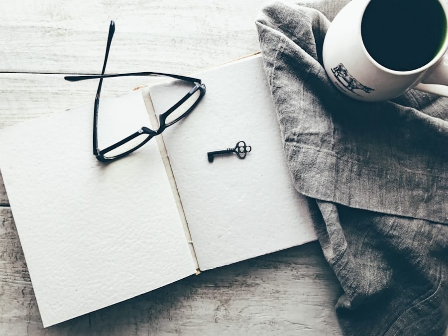Overhead view of an open blank notebook with black glasses and a small vintage key on its pages, next to a white coffee mug on gray linen atop a rustic wooden table.