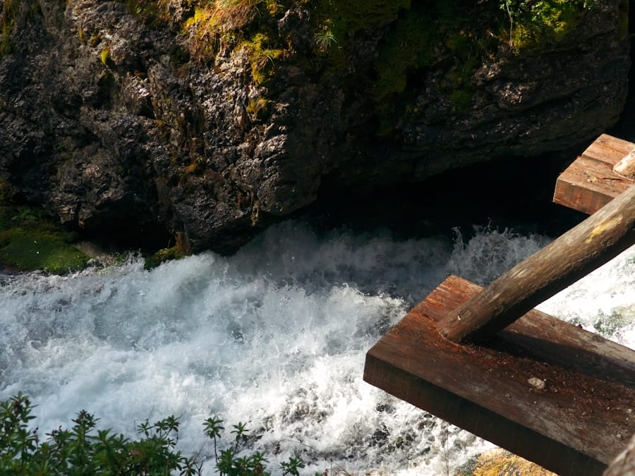 Whitewater rushes through a narrow rocky gorge beneath a moss-covered cliff, viewed from a wooden platform in the foreground.